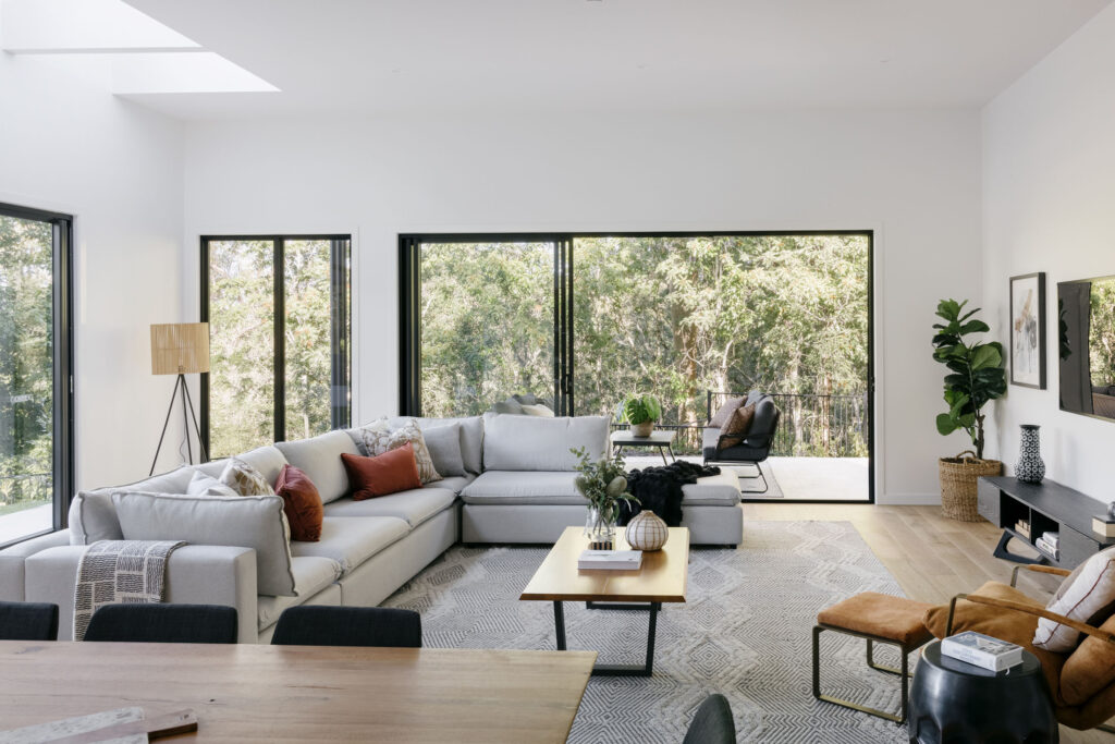 The image features a high ceiling living room with bush backdrop and plenty of natural light and space.